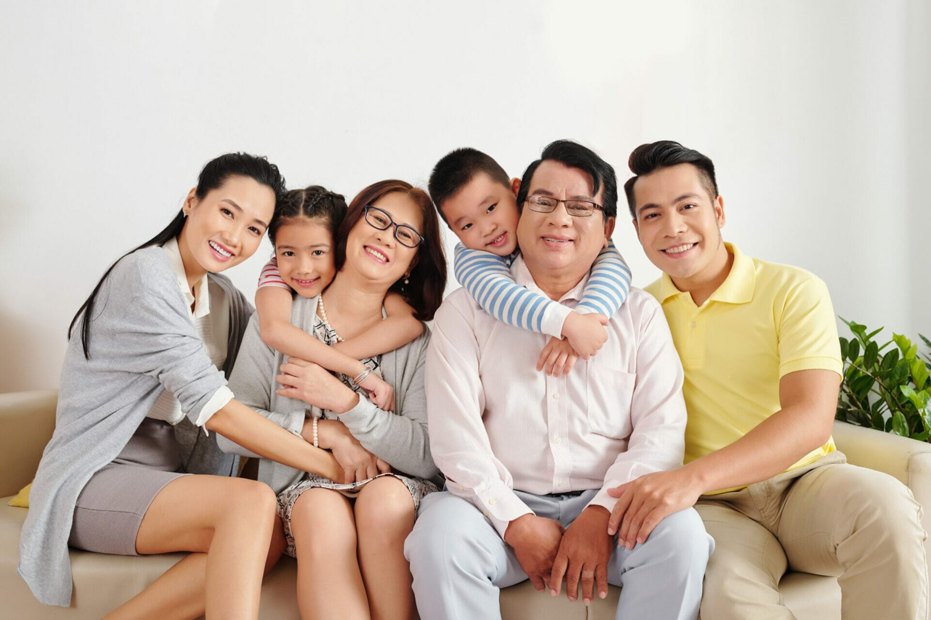 Cheerful Vietnamese children, parents and grandparents sitting on sofa in living room, hugging and looking at camera