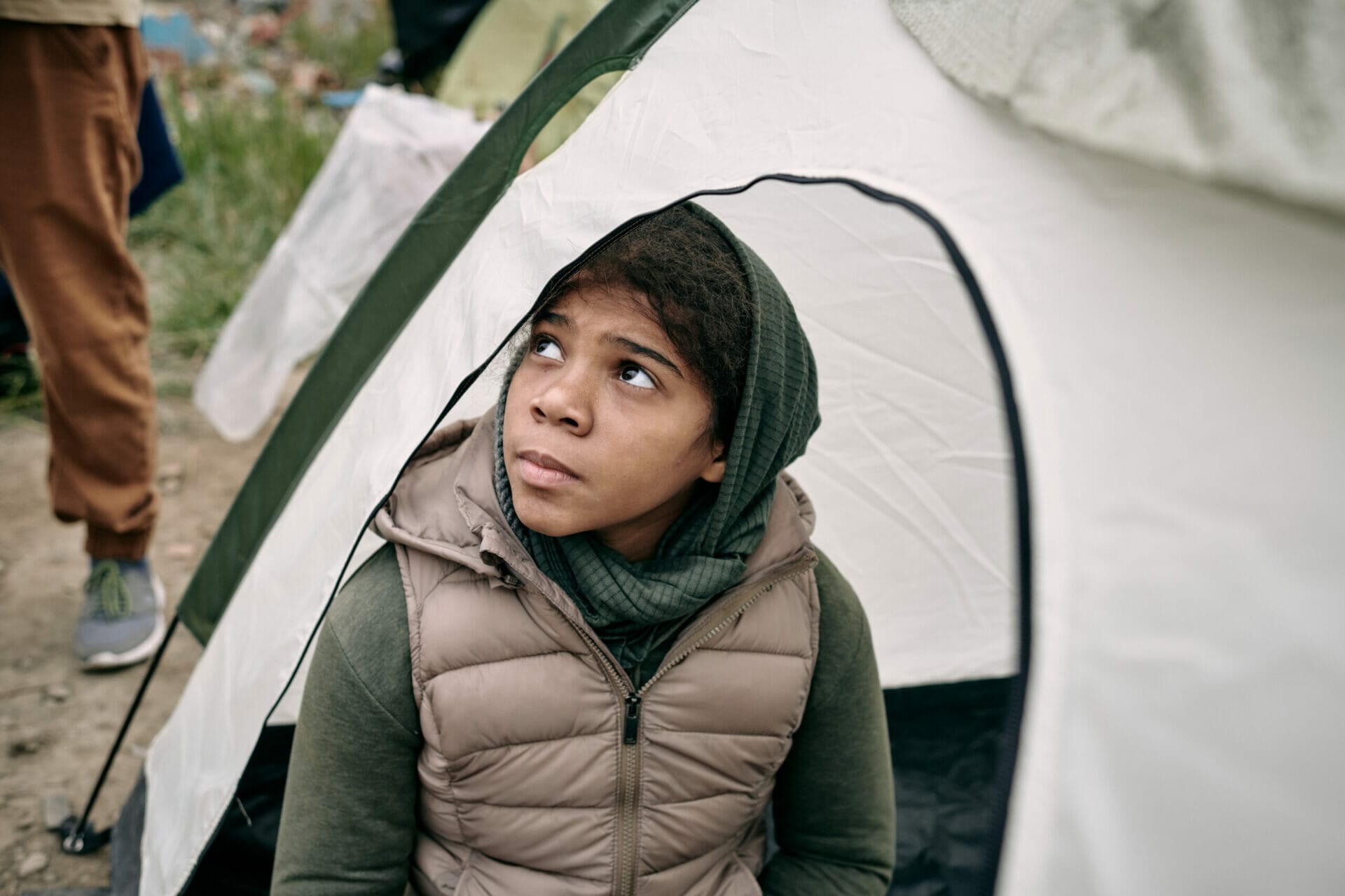 Serious homeless refugee middle-eastern girl in headscarf and vest sitting inside of tent and looking up hopefully